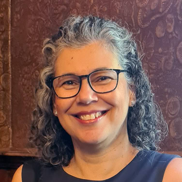 Headshot of Rose May smiling with shoulder length curly hair and glasses.